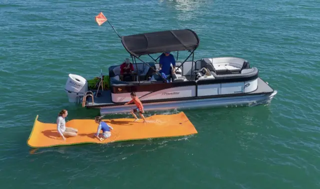 Pontoon boat anchored near a sandbar with kids playing on an orange floating mat